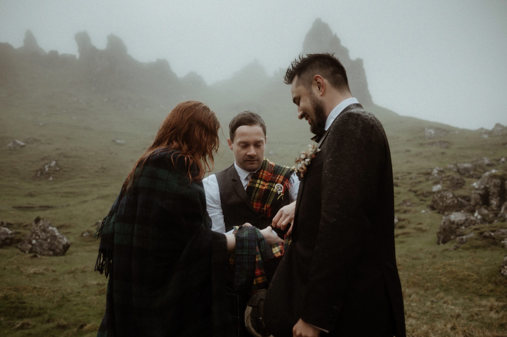 A traditional Scottish hand-fasting taking place during a wedding in Scotland outdoors in the rain with Bride and groom holding tartan ribbon near the Old man of Storr on Skye