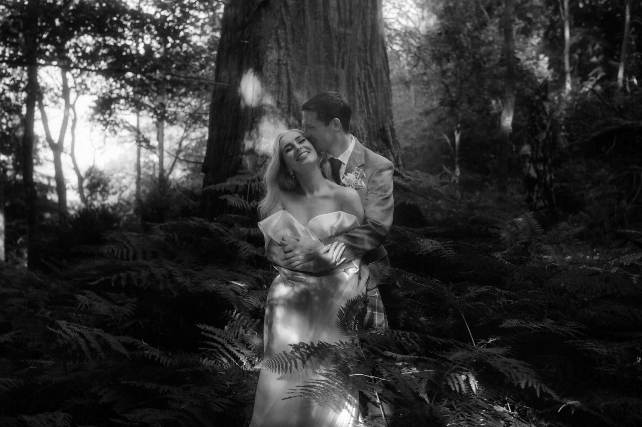bride and groom wandering in woodland and fern hugging and kissing and laughing during their wedding in Scotand at Birkhill house