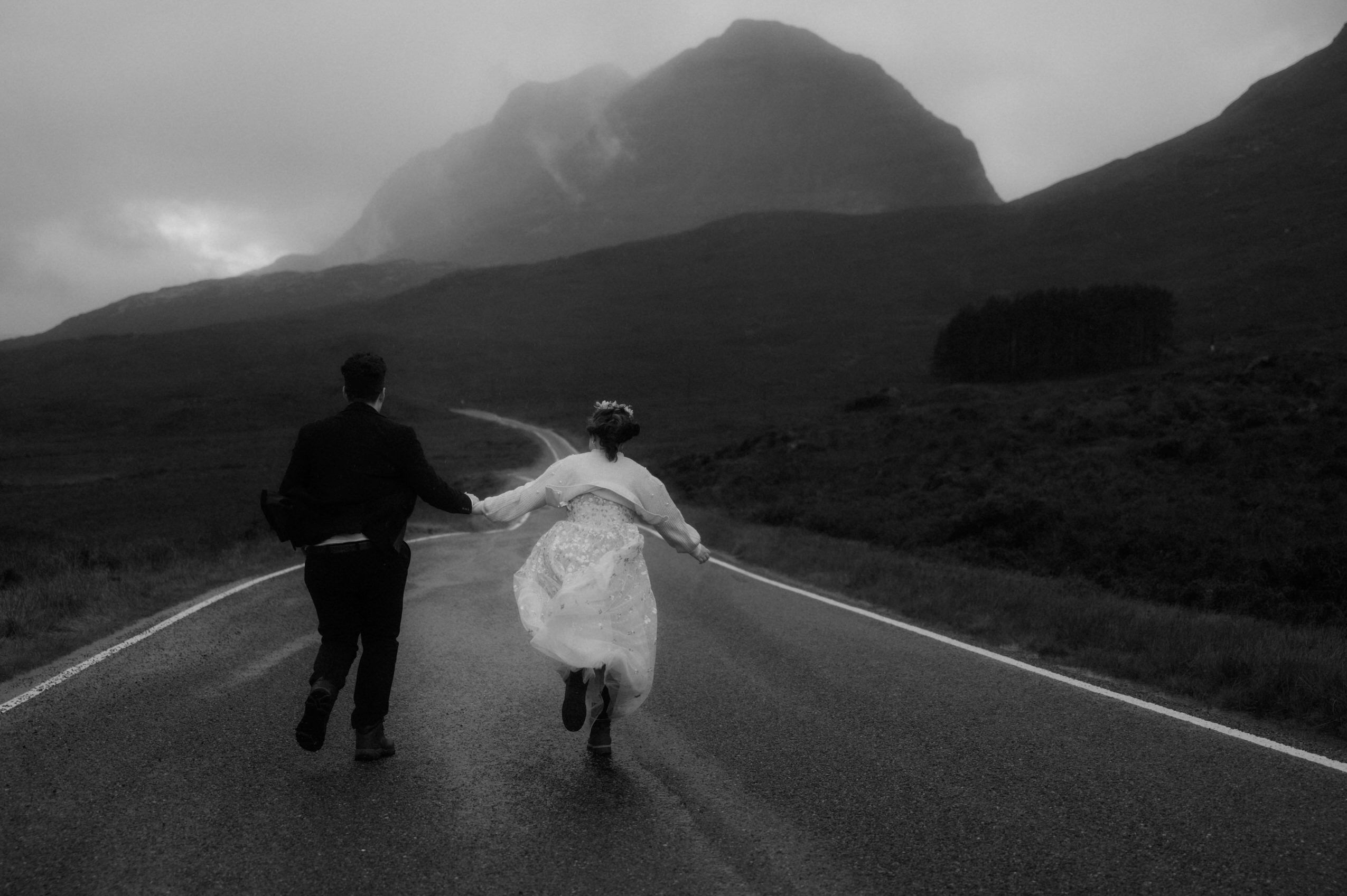 couple running in the mountains during their Torridon elopement with the liathach mountain in the background and the rain