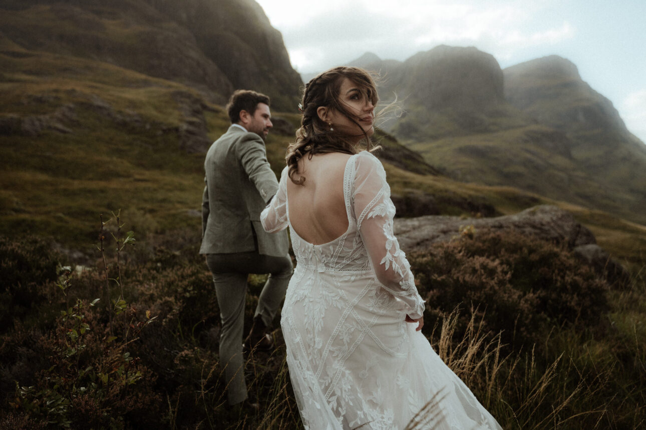 elope to Glencoe couple walking in the mountains with bride looking over her shoulder as husband leads on at the study in Glencoe valley in scotland
