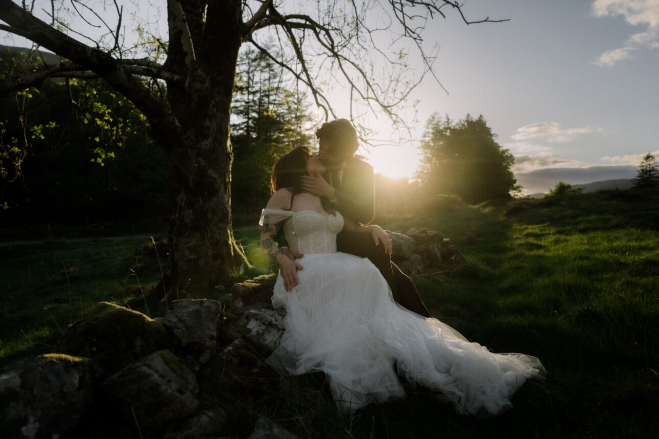 elope to scotland couple sit under a tree in Glencoe kissing with the sun whinging brightly behind them in a romantic way