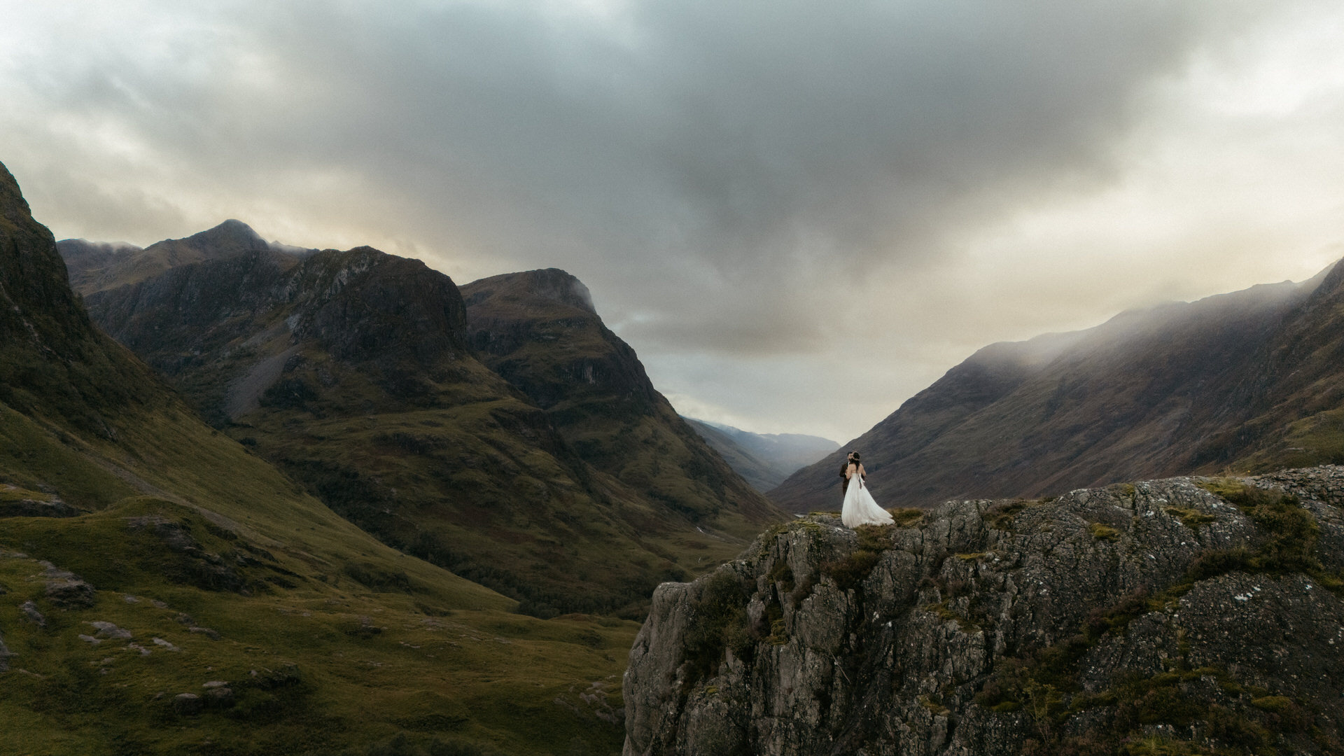 a drone shot of a couple who eloped to glance in scotland and are standing in th mountains